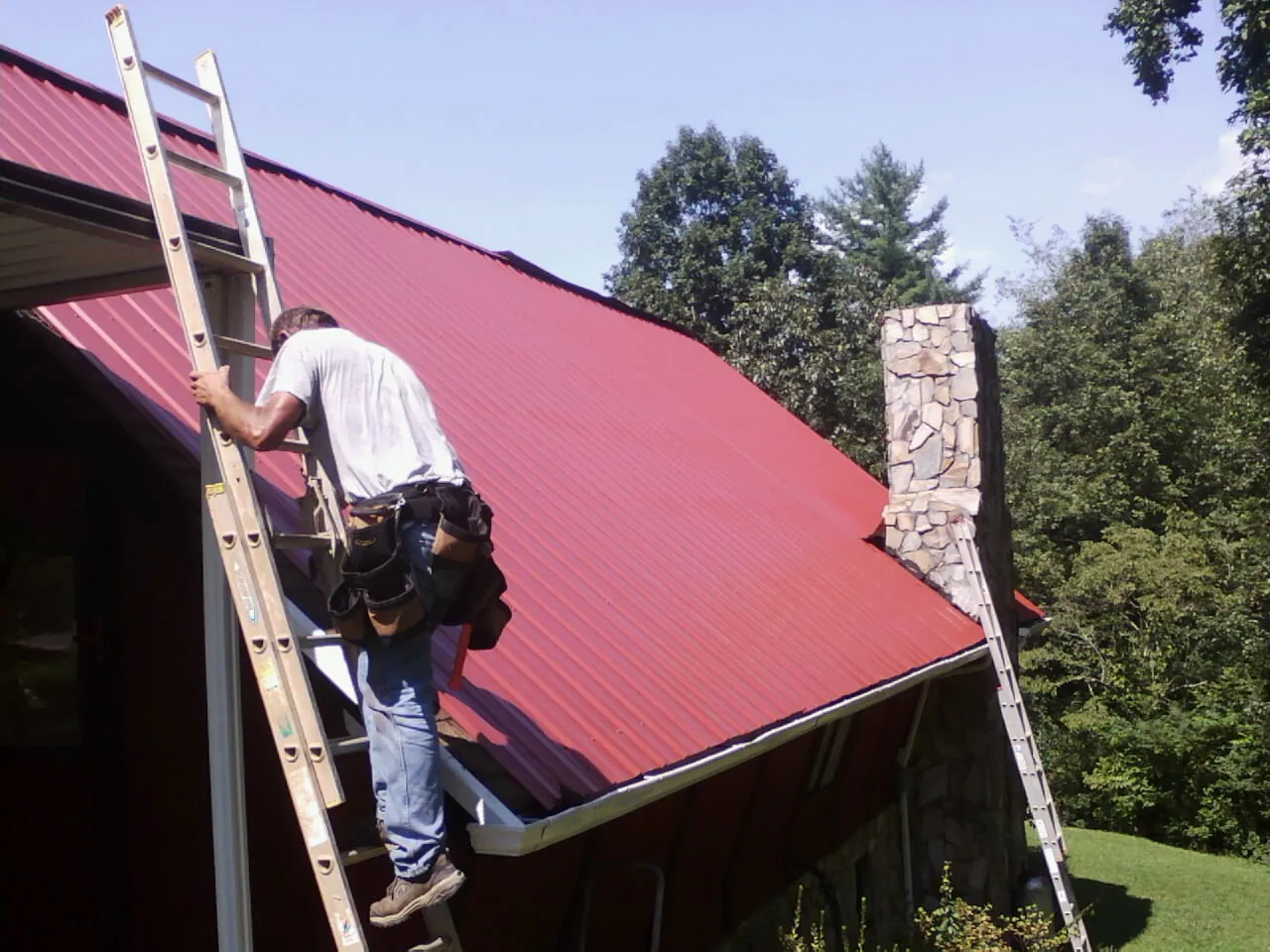 Roofer on a ladder beside a red metal roof and stone chimney — Soffit & Fascia Repair by Roofing Brothers in Hampton